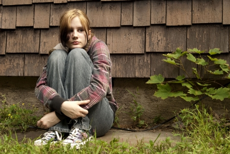 5907015 - a teenage girl with a sad expression sits against a run-down house.